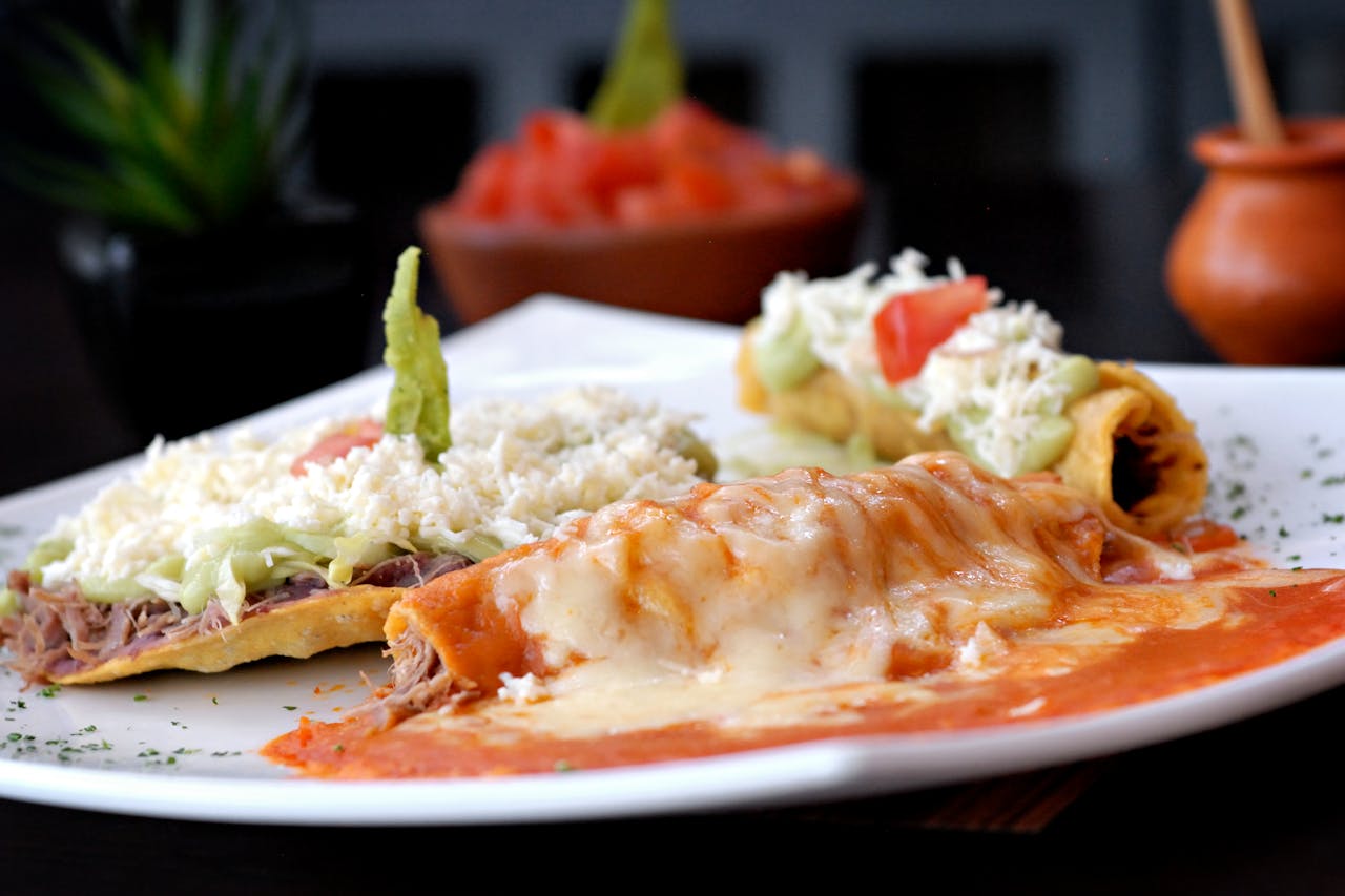 about-02 Mouth-watering Mexican enchiladas and tacos garnished with cheese and avocado on a white plate.